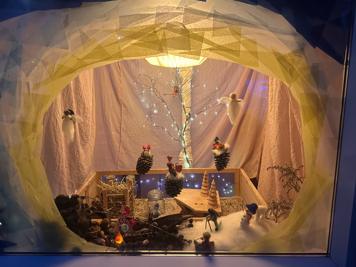 Een sfeervol verlicht tafereeltje gemaakt van doorzichtig papier, takjes, dennenappels en vilten kerstcreaties